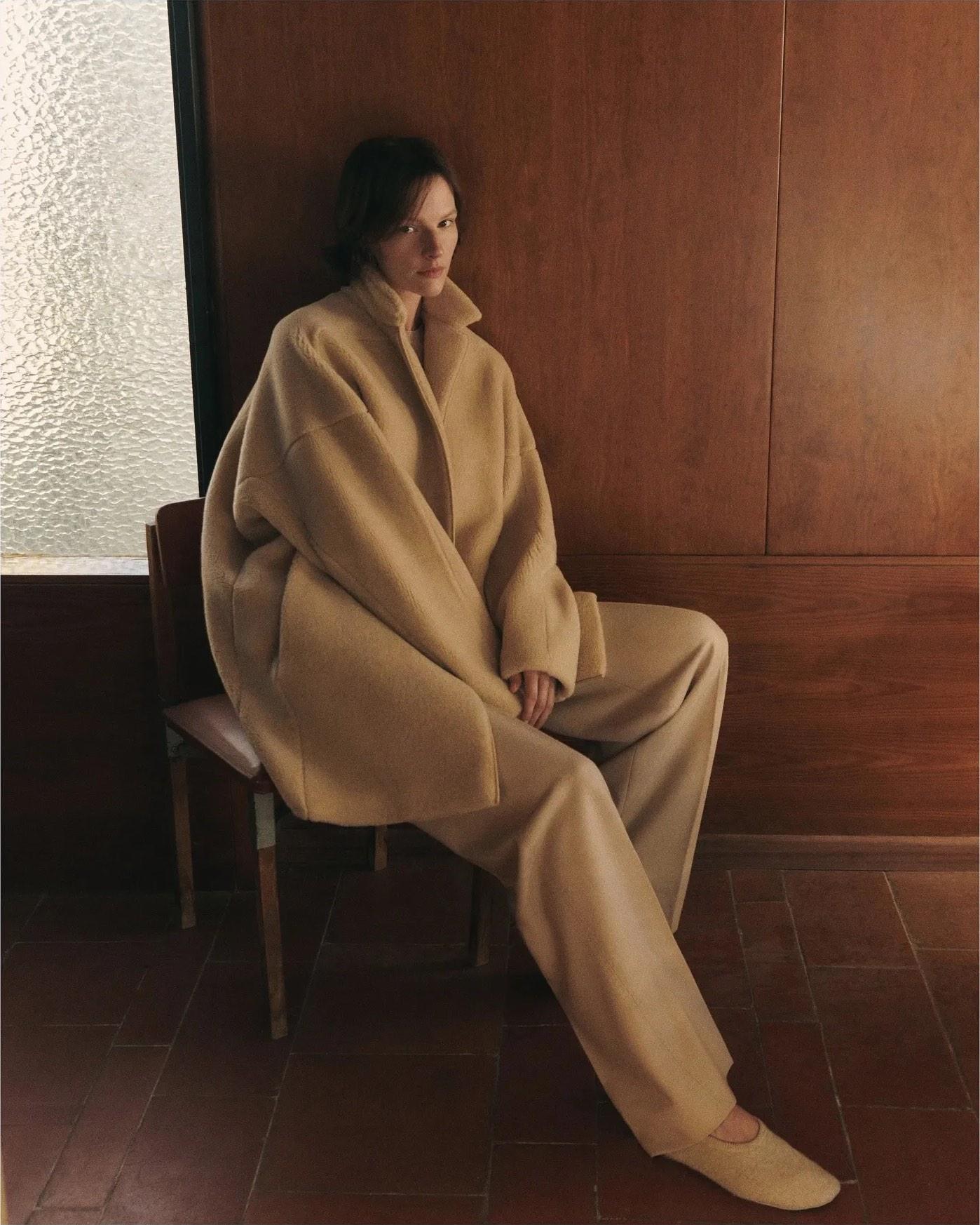 A model seated against wood panelling in an oversized camel coat and matching trousers