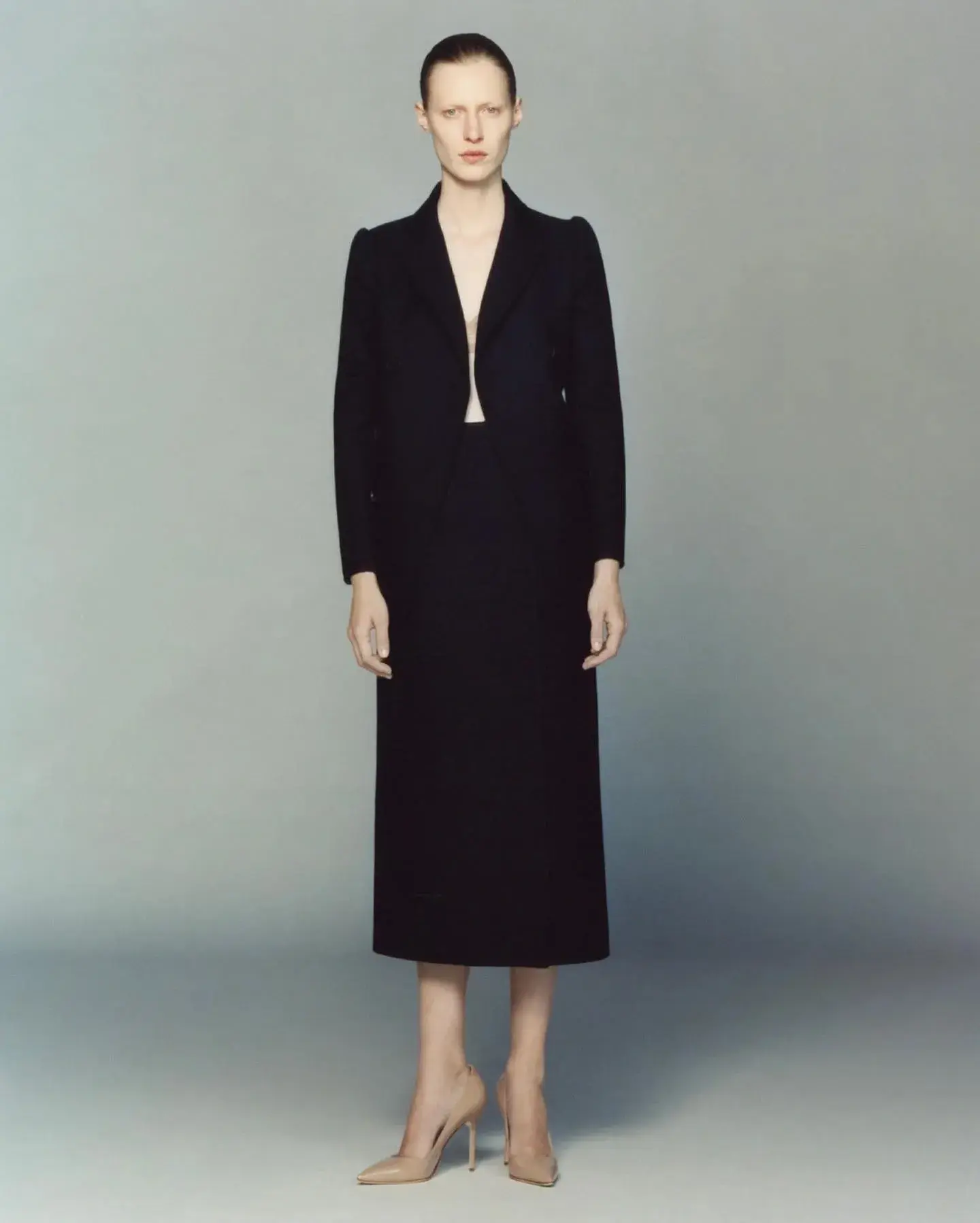 A model in a long black tailored coat against a grey studio wall
