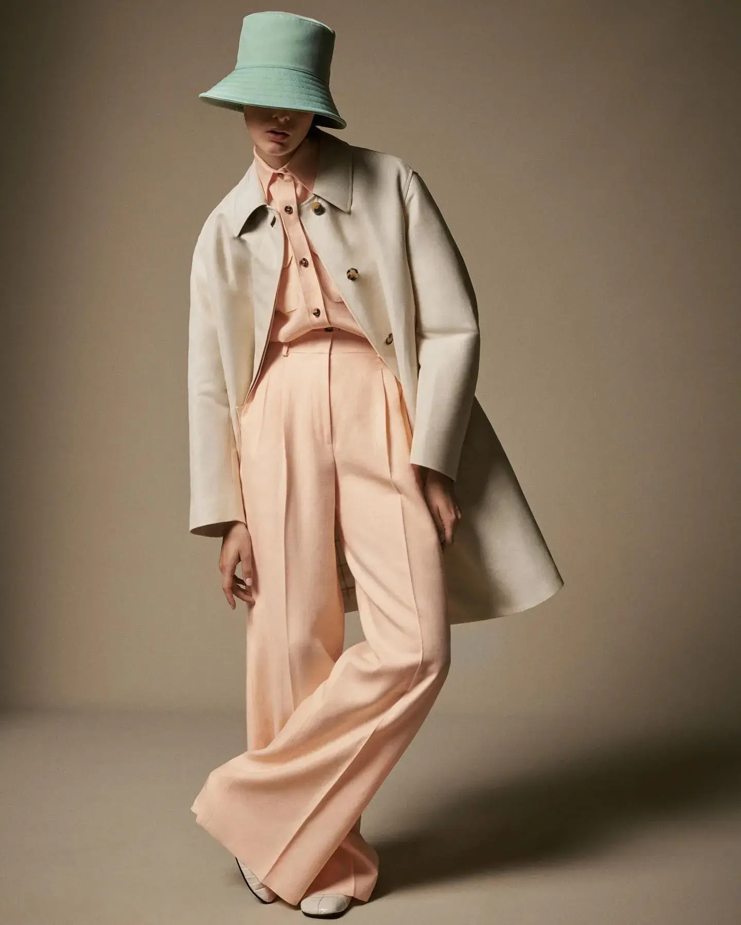 A model in a mint bucket hat and cream coat over peach wide-leg trousers
